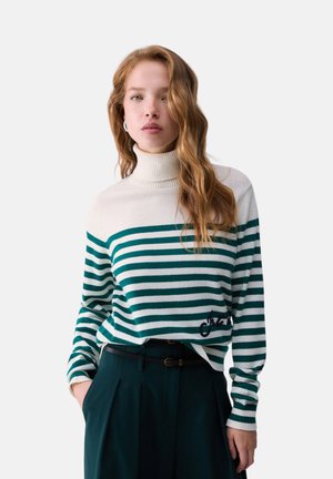Donna con capelli rossi ondulati che indossa un maglione bianco a collo alto con strisce orizzontali verdi e pantaloni scuri a vita alta con cintura, in piedi con una mano in tasca.