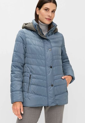 Frau trägt eine hellblaue gesteppte Jacke mit vorderen Druckknöpfen, Reißverschlusstaschen, grauem Kapuzenfutter und einem grauen Rollkragenpullover.