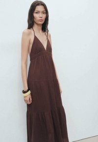Robe midi brune en tissu léger et texturé. Avec un décolleté en V, des bretelles fines et un design à volants. Accentuée par des bracelets.
