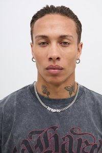 T-shirt gráfica preta com texto vermelho, colar de corrente prateado e tatuagens visíveis de uma rosa e uma borboleta. Retrato em close-up.