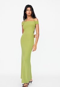 Robe maxi verte, moulante, avec un décolleté carré, des manches dénudées et une texture lisse. Portée avec des talons noirs à lanières.