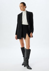 Blazer nero su un maglione bianco a collo alto, abbinato a una gonna nera con rifiniture in pizzo e stivali alti con tacco.