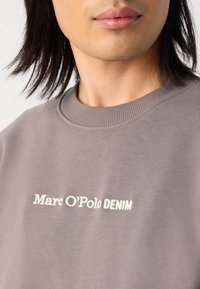 Pelēks džemperis no mīksta auduma, ar apaļu kakla izgriezumu un izšūtu "Marc O'Polo DENIM" logo baltā krāsā. Gluda tekstūra un minimālisma dizains.