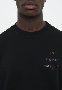 Sudadera negra con cuello de canalé, que presenta un texto bordado multicolor "PS Paul Smith" en la parte frontal. Textura suave, diseño informal.