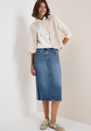 Vrouw die glimlacht, gekleed in een beige hoodie, een beige knoopvest van fleece, een blauwe denim midi-rok en beige sneakers, staand tegen een lichte muur.