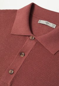 Polo couleur rouille avec une surface texturée, un col classique et trois boutons bruns à l'avant. Étiquette visible à l'intérieur du col.