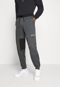 Grå sweatpants med en svart panel, elastiskt midjeband med dragsko och en gul "adidas"-logotyp. Texturerad tyg och avsmalnande design.