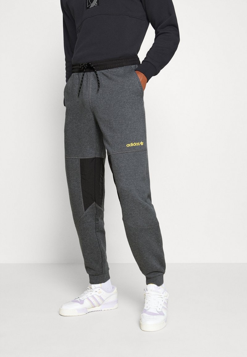 Grå sweatpants med en svart panel, elastiskt midjeband med dragsko och en gul "adidas"-logotyp. Texturerad tyg och avsmalnande design.