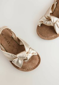 Guldmetalliska platta sandaler med en knuten frontdesign, mjukt mockainlägg och sömnadsdetaljer runt kanten.