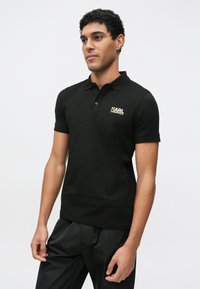 Črna polo majica iz bombaža z ovratnikom, kratkimi rokavi in zlatim logotipom na prsih. Oblečena s črnimi, širokimi hlačami.