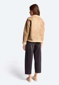 Hnědá semišová bunda s beránkovým lemem, oversized střih, na světlém pozadí, v kombinaci s tmavými kalhotami širokého střihu a sandály.