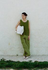 Groene geribbelde gebreide tanktop, textuur olive cargobroek en een lichtgekleurde tas. De vrouw staat tegen een witte muur met een grasachtige ondergrond.