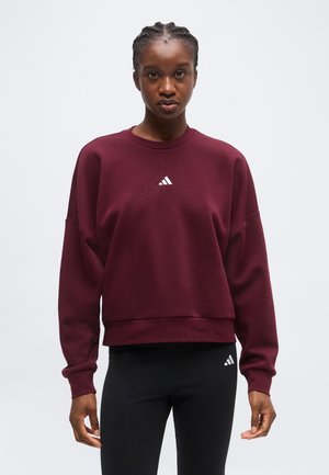 Mujer con cabello trenzado, vestida con una sudadera burdeos y mallas negras con pequeños logos blancos de Adidas, está de pie mirando hacia adelante.