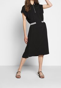 Black dress with short sleeves, zip front, and white striped waistband. Features button details at the side and a mid-length hem.