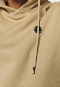 Felpa beige con cappuccio e cordino, caratterizzata da una toppa rotonda nera con un logo. Il tessuto è morbido e strutturato.