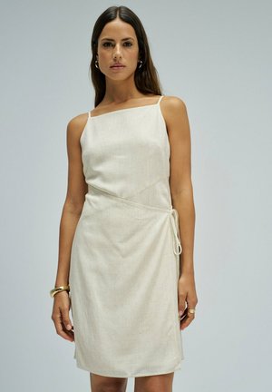 Mujer que lleva un vestido cruzado beige sin mangas con tirantes finos, pendientes de aro dorados, pulsera y anillo, de pie frente a un fondo gris claro.