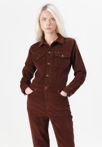 Bruine corduroy jumpsuit met een aansluitende uitstraling, knoopsluiting aan de voorkant, twee borstzakken en zijzakken. Gestructureerde stof met een zachte afwerking.