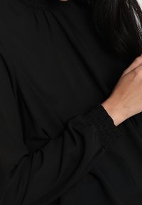 Schwarze Bluse mit hohem, gerafftem Kragen. Lange, lockere Ärmel mit elastischen Bündchen. Weicher, glatter Stoff, minimalistisches Design.