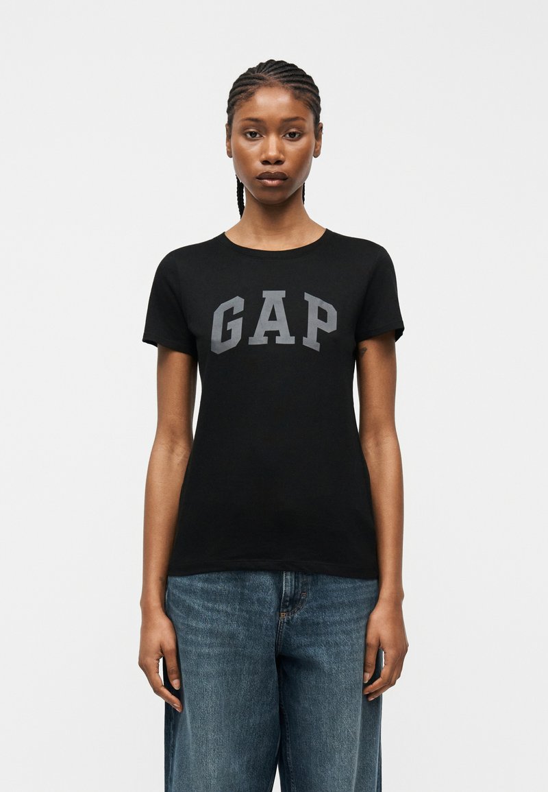 Jeune femme aux cheveux tressés portant un t-shirt noir GAP et un jean bleu ample, debout devant un fond blanc uni.