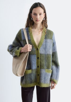 Jeune femme portant un cardigan à carreaux verts et bleus, tenant un sac à main beige avec un pantalon ample foncé sur un fond clair uni.