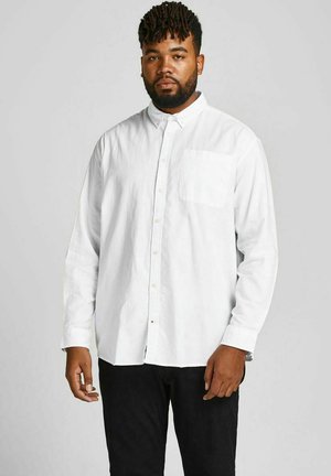 Chemise blanche à manches longues avec un poche sur la poitrine, fabriquée dans un tissu texturé. Associée à un pantalon noir, sur un fond neutre.