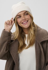 Witte fleece muts met een opgerolde boord, gedragen met een bruine wollen jas over een crème trui. De achtergrond is eenvoudig en licht.