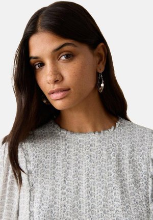 Hellgrauer, strukturierter Pullover mit wellenförmigem Saum, präsentiert mit gemustertem Stoff und weichem, geschwungenem Ausschnitt. Model trägt hängende Ohrringe.