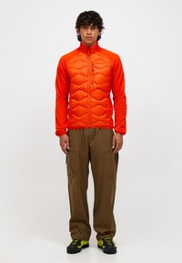 Giacca imbottita arancione con texture trapuntata e colletto alto, abbinata a pantaloni cargo marroni con tasche e scarpe da escursionismo nere con accenti al neon.