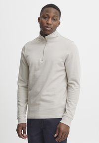 Light beige zip-up pullover with long sleeves and a high collar, made from soft fabric, featuring a central zipper and ribbed cuffs.