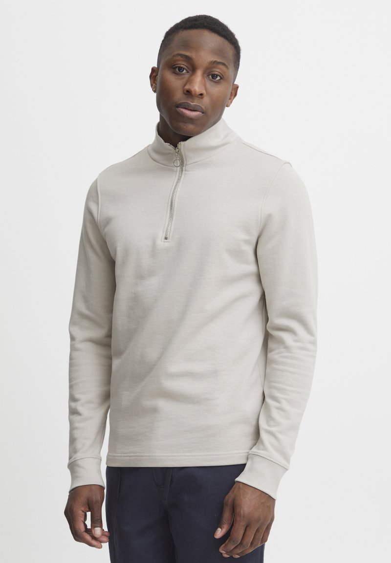 Light beige zip-up pullover with long sleeves and a high collar, made from soft fabric, featuring a central zipper and ribbed cuffs.