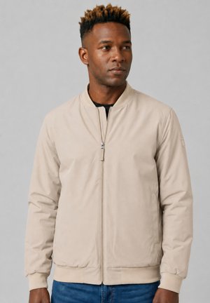 Uomo con capelli corti e ricci che indossa una giacca beige con zip e jeans blu, che guarda leggermente verso destra su uno sfondo grigio uniforme.