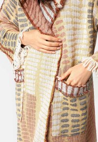 Textúrovaný šál v žltých, béžových a ružových vzoroch. Má strapce na okrajoch a dve bočné vrecká. Materiál vyzerá mäkko a vrstvené.