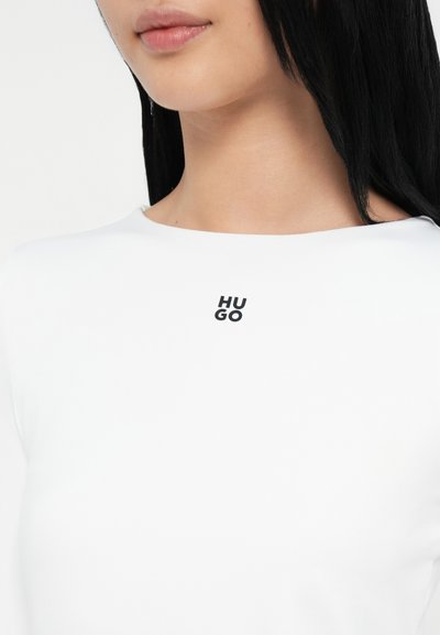 Бяла тениска с кръгло деколте и къси ръкави, с малко черно лого "HU" и "GO" в централната част на гърдите.