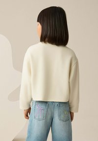 Cream sweatshirt with a loose fit and three-quarter sleeves, paired with light blue jeans featuring embroidered pink and green accents on the back pockets.