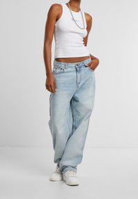 Person wearing a white sleeveless top, silver chain necklace, light blue loose jeans, and white sneakers with one hand in pocket.