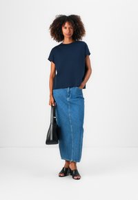 Marineblå t-skjorte med korte ermer kombinert med et denim maxi-skjørt, svarte sandaler med åpen tå og en liten svart håndveske, satt mot en hvit bakgrunn.