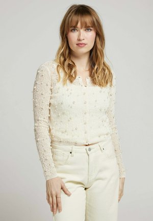 Top court en tricot ivoire avec des embellissements en forme de perles, manches longues et une rangée de boutons au niveau du col, associé à un jean blanc taille haute.