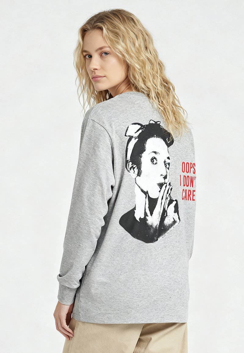 Jeune femme aux cheveux blonds portant un sweat-shirt gris avec un graphisme en noir et blanc d'une femme surprise et un texte rouge disant « OUPS, JE M’EN FOUS ».