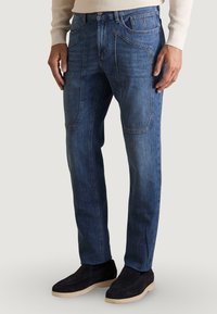 Vaqueros de mezclilla azul oscuro de corte recto, con múltiples bolsillos, acentos de costura clara y una textura suave. Combinados con zapatos negros.