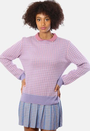 Donna con capelli ricci che indossa un maglione a quadretti rosa e lavanda e una gonna a pieghe blu a quadretti, in piedi con le mani sui fianchi.