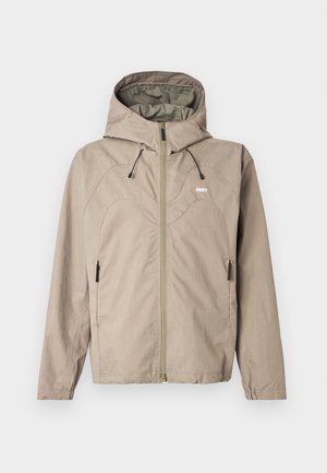 Μπεζ ζακέτα με φερμουάρ και κουκούλα, με καμπύλη ραφή στο σχέδιο, ρυθμιζόμενα κορδόνια και πλαϊνές τσέπες με φερμουάρ. Το λογότυπο εμφανίζεται στο στήθος.