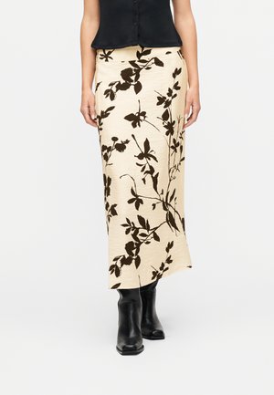 Naine, kes kannab kreemjat midi-seelikut musta lillemustriga, musta varrukateta nööpidega topi ja musti saabastest pahkluutel, seisab valgel taustal.