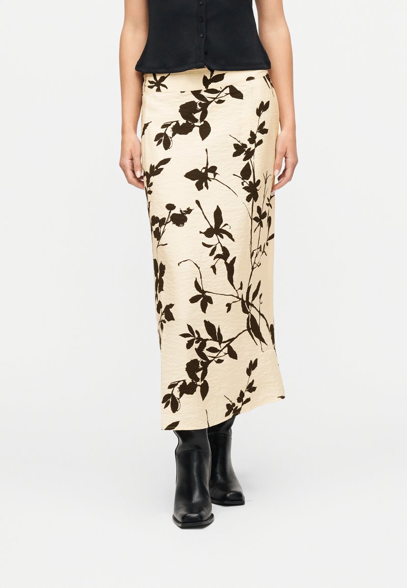 Femme portant une jupe midi crème à imprimé floral noir, un haut noir sans manches boutonné, et des bottines noires, debout sur fond blanc.