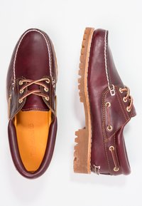 Chaussures en cuir bourgogne avec semelles en caoutchouc marron, bout rond, couture blanche et œillets dorés. L'intérieur présente une doublure jaune.