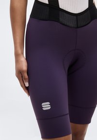 Nahaufnahme einer Person, die dunkelviolette, gepolsterte Fahrradhosen mit einem schwarzen Bund und einem weißen Logo am linken Oberschenkel trägt.