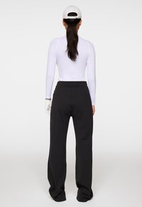 Kvinna som bär vit keps, vit långärmad topp, svarta vida byxor och en vit handske, stående vänd bort från kameran mot en neutral bakgrund.