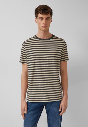 Młody mężczyzna stoi, ubrany w beżowo-czarną, poziomo paskowaną koszulkę z krótkim rękawem i niebieskie dżinsy, na jednolitym szarym tle.