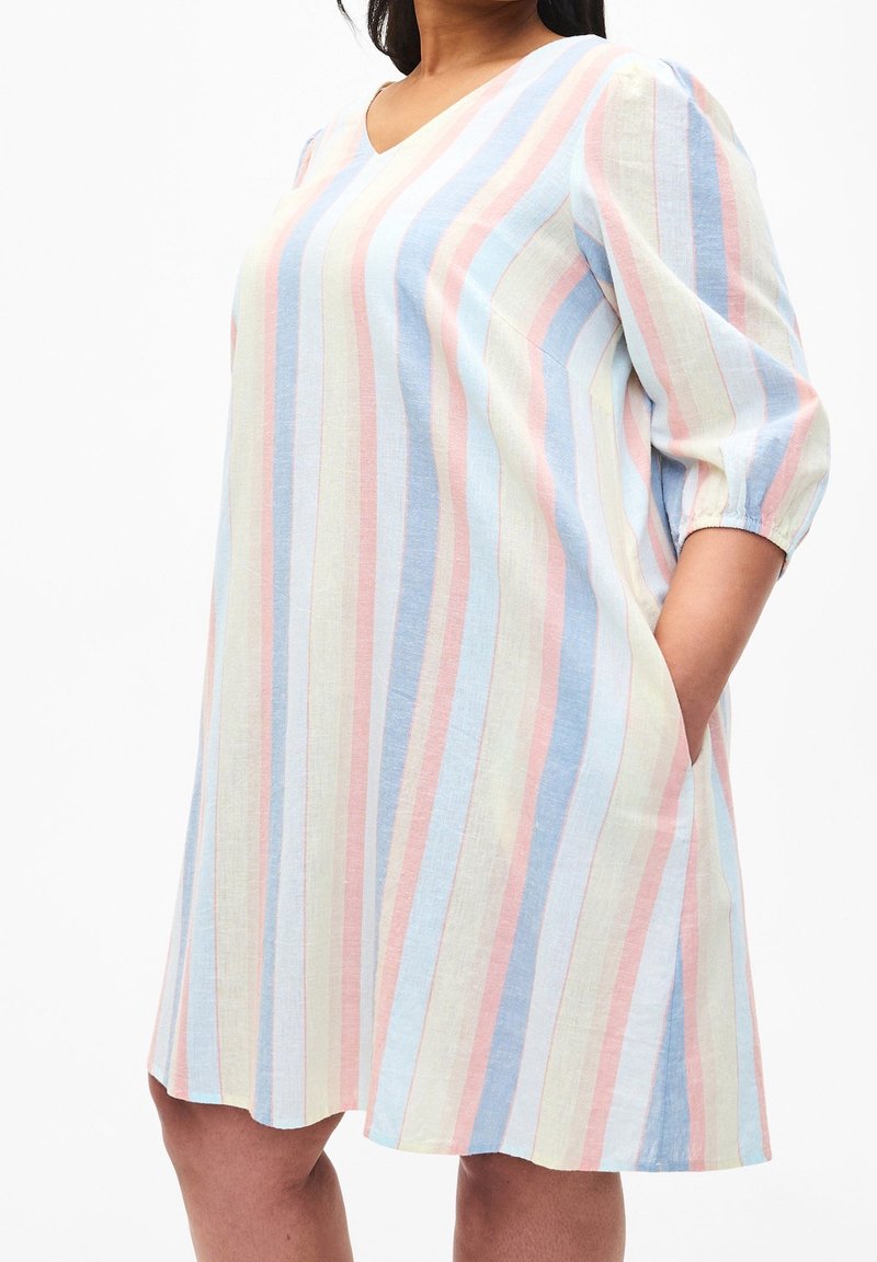 Femme portant une robe ample mi-longue avec des rayures verticales pastel et des manches trois-quarts bouffantes, main dans la poche.