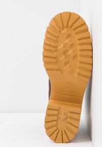 Hnědá podrážka boty s hlubokými vzory, s vyvýšeným logem Timberland a světlým hnědým gumovým materiálem. Lehce nakloněná na bílém pozadí.