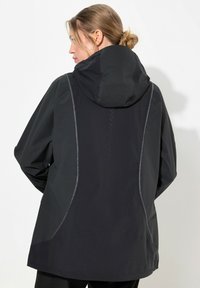 Chaqueta negra impermeable con capucha, que presenta costuras grises en contraste y un corte relajado. Textura suave y diseño minimalista.